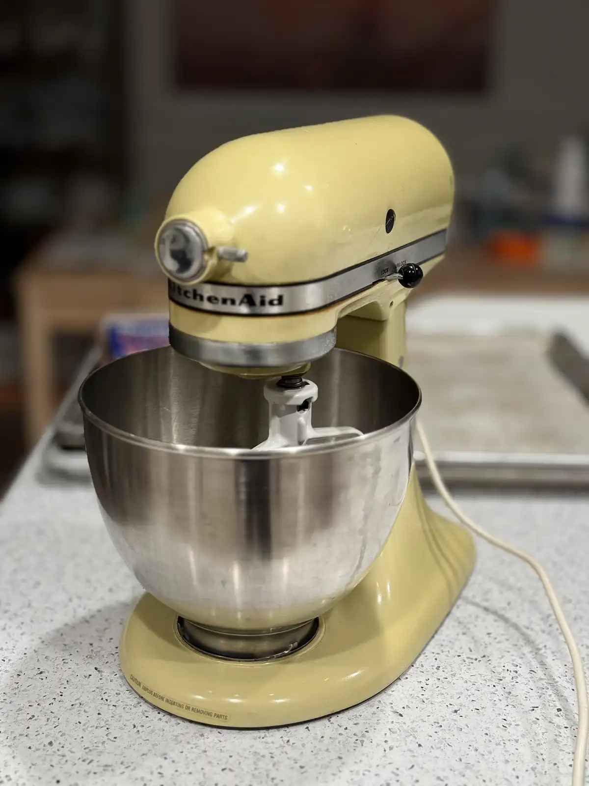 A pale yellow KitchenAid stand mixer sits on a speckled countertop with its stainless steel bowl and paddle attachment…