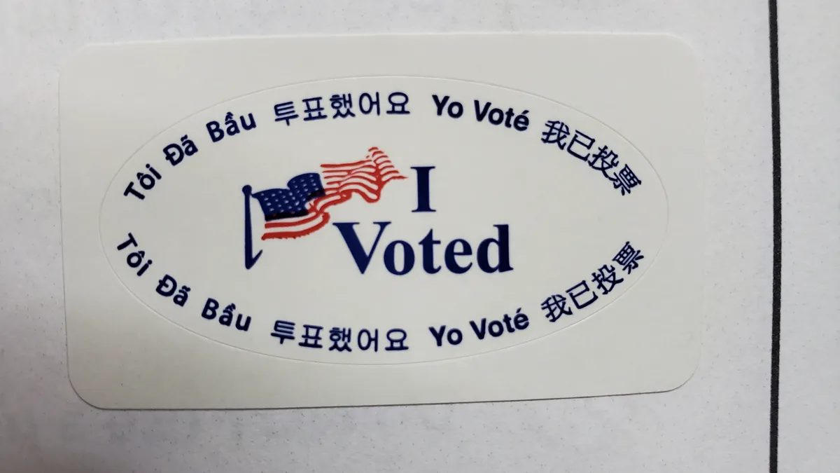 A close-up of a white oval 'I Voted' sticker featuring a small American flag graphic and the phrase 'I Voted' in large blue…