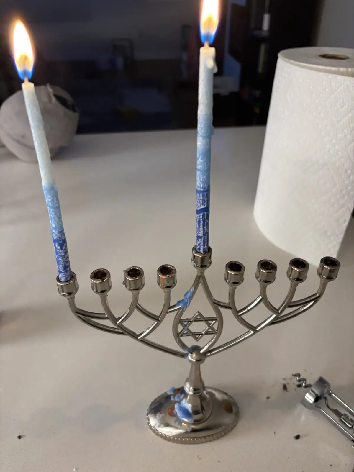 A silver menorah with two lit blue-and-white candles stands on a white countertop, with melted wax stains on its base and…