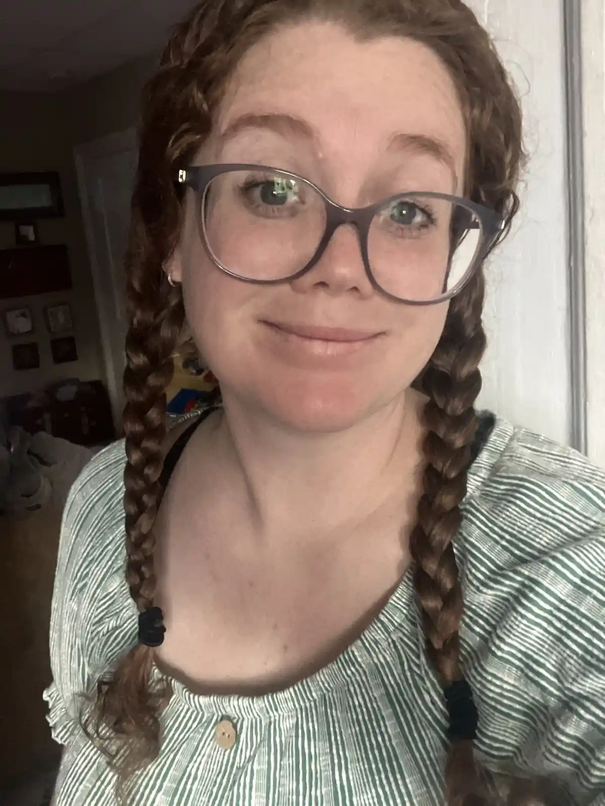 A person with reddish-brown hair styled in two braids and wearing large, dark-rimmed glasses smiles slightly at the camera…