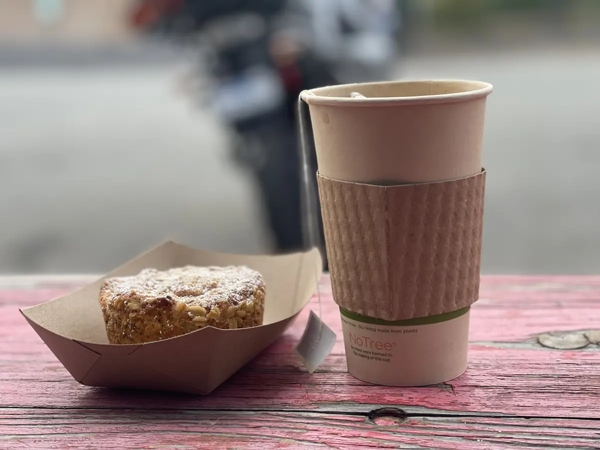 A close-up photo shows a paper cup with a 'NoTree' sleeve and a tea bag beside a pastry in a paper boat, both resting on a…