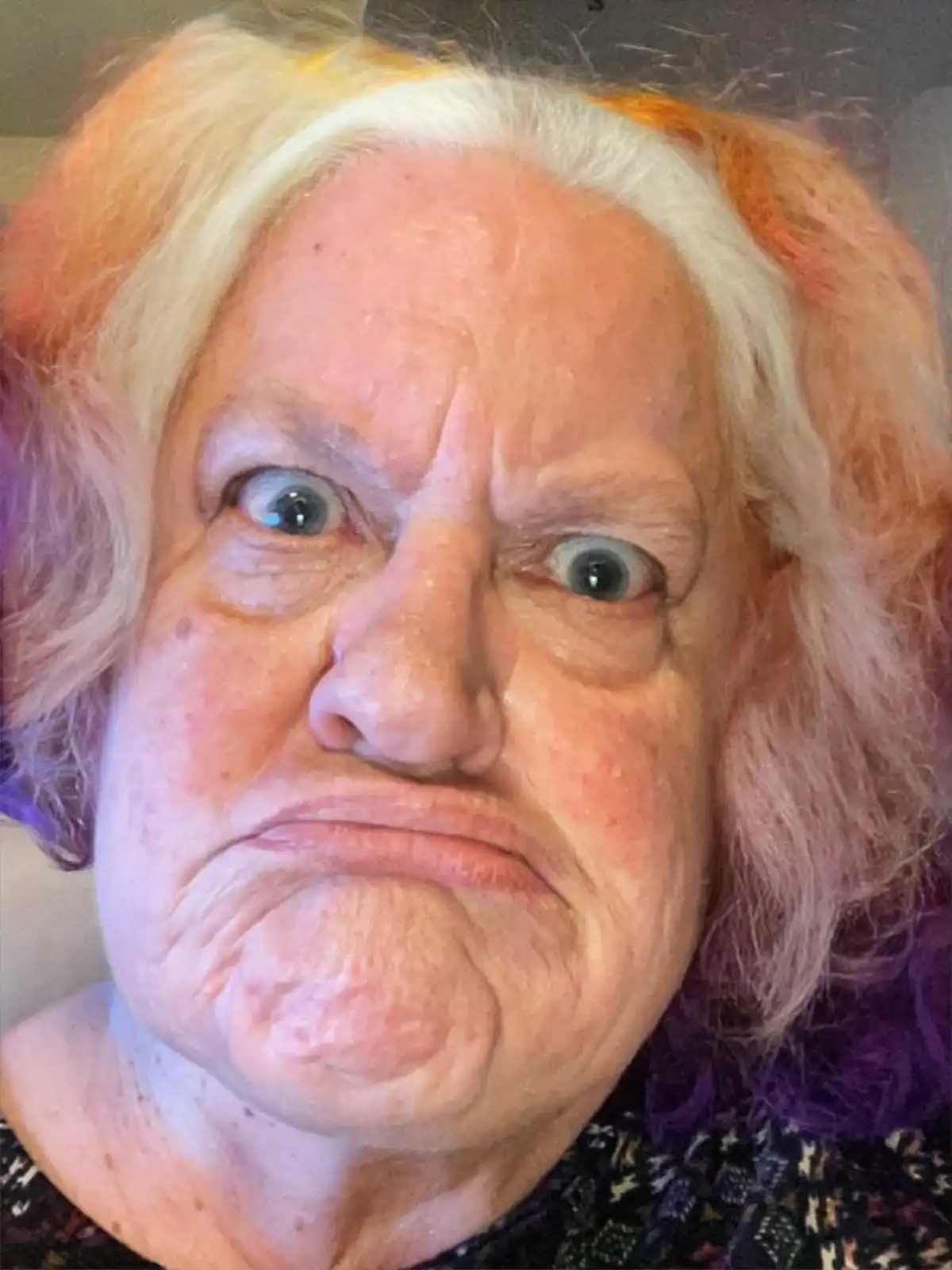 A close-up photo of an older woman with white and orange-pink hair, making a comically exaggerated angry face with wide eyes…