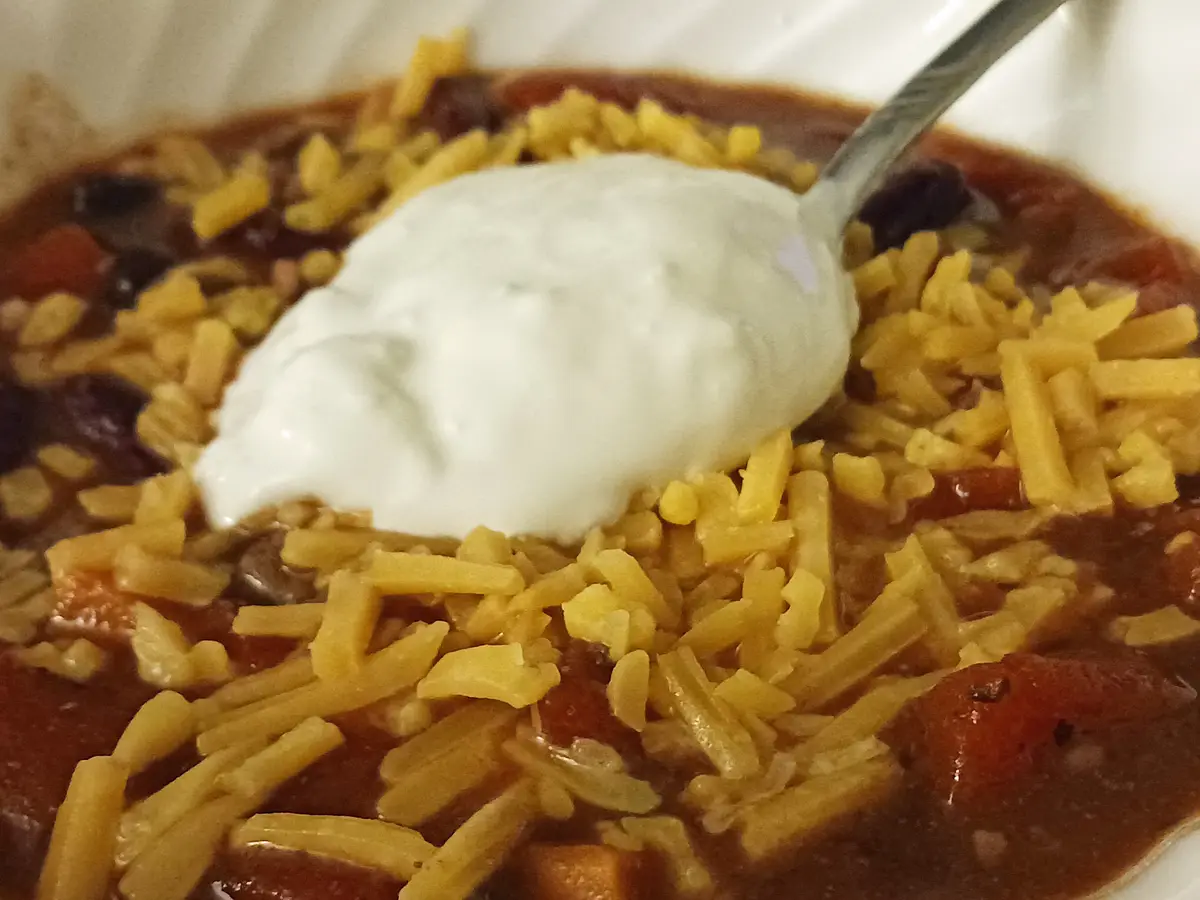 A close-up photo shows a bowl of chili topped with shredded cheese and a large dollop of sour cream, with a spoon resting in…