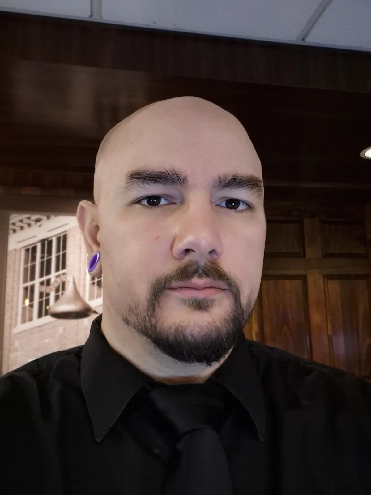 A bald man with a goatee and a purple earring looks directly at the camera with a neutral expression while wearing a black…