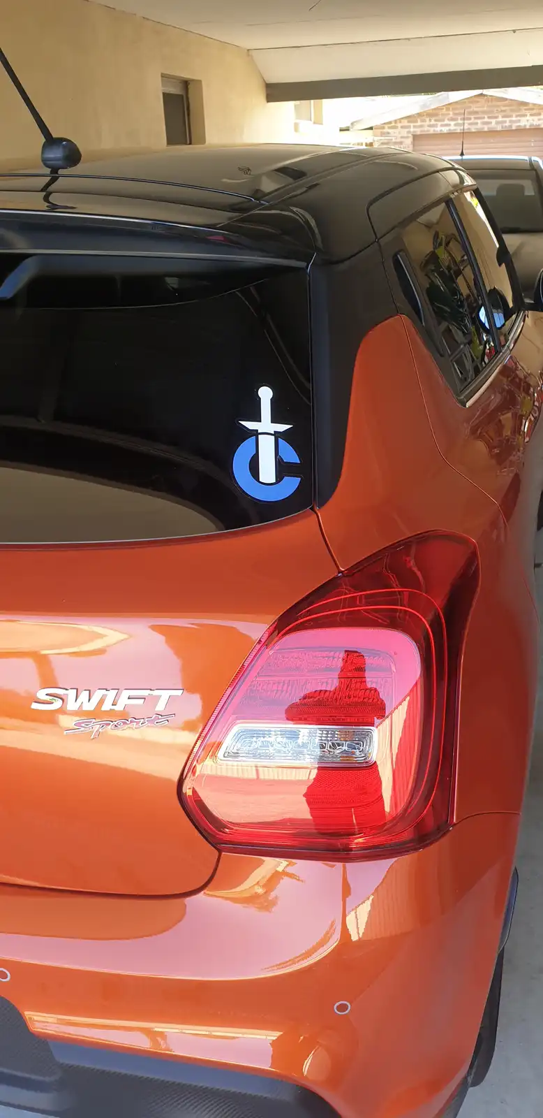 The rear of an orange Suzuki Swift Sport car is shown, featuring a 'Swift Sport' badge and a custom white…