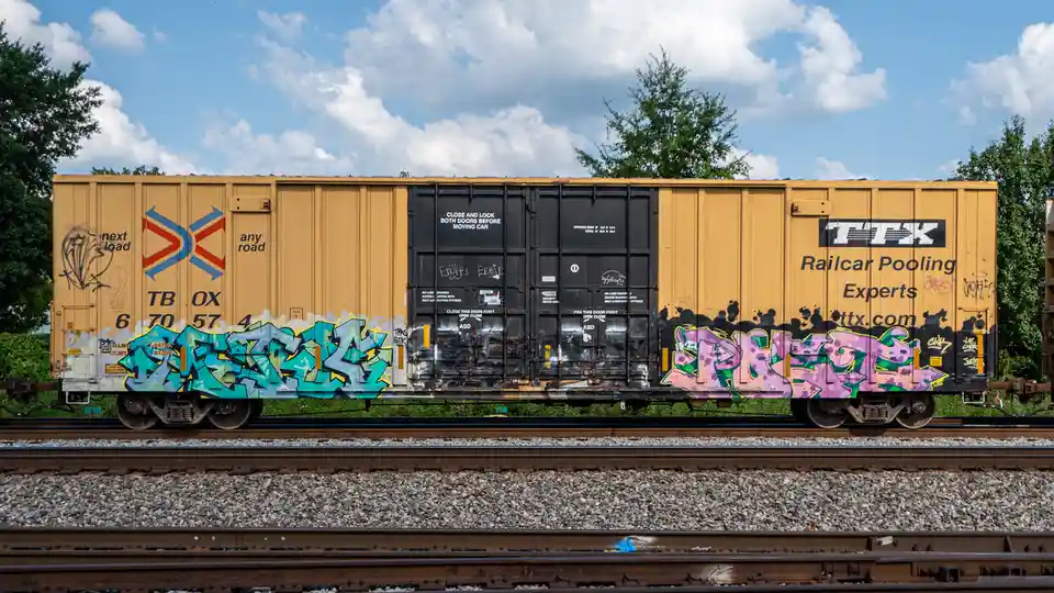 A yellow freight train car sits on tracks under a partly cloudy sky, covered in colorful graffiti on its sides and doors…