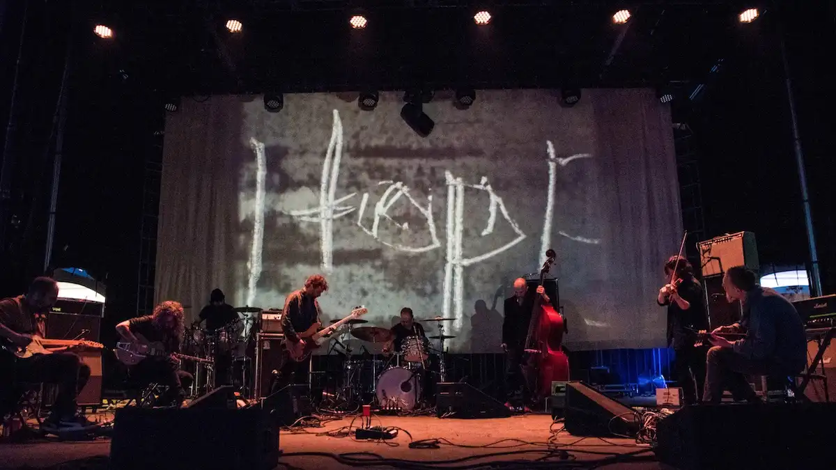 A live band performs on a dimly lit stage with a large backdrop screen projecting the word 'hope' in a rough, handwritten…