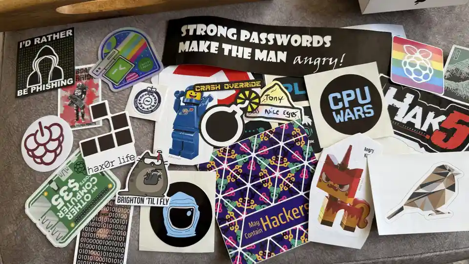 A collection of tech and hacker-themed stickers scattered on a gray surface, including a large black sticker with white text…