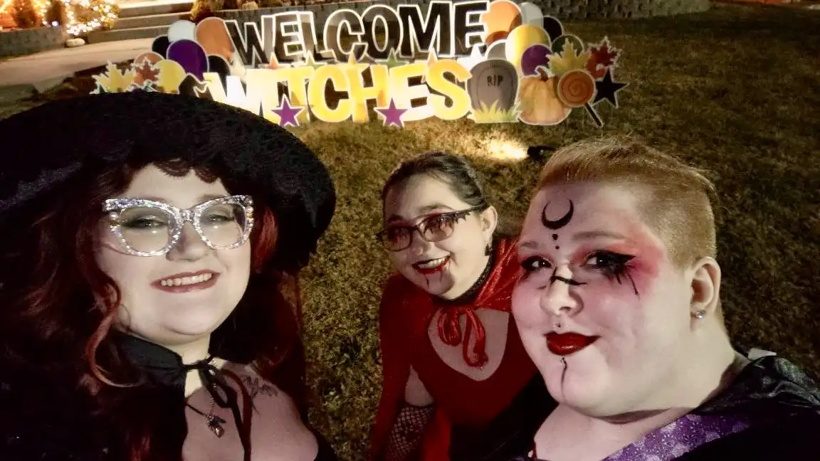 Three women in Halloween witch costumes pose for a photo in front of a decorative sign that reads 'WELCOME WITCHES'. The…