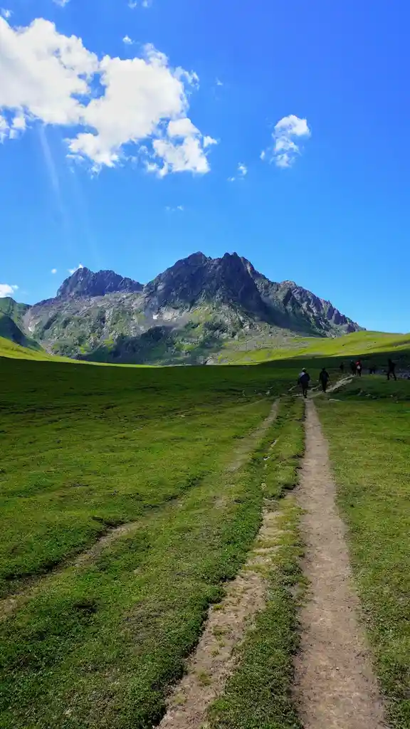 A scenic mountain landscape with a dirt trail cutting through vibrant green grass, leading toward a rugged, rocky peak under…