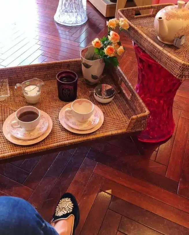 A first-person view shows a person's feet in ornate black slippers resting on a wooden floor, looking up at a wicker tray…