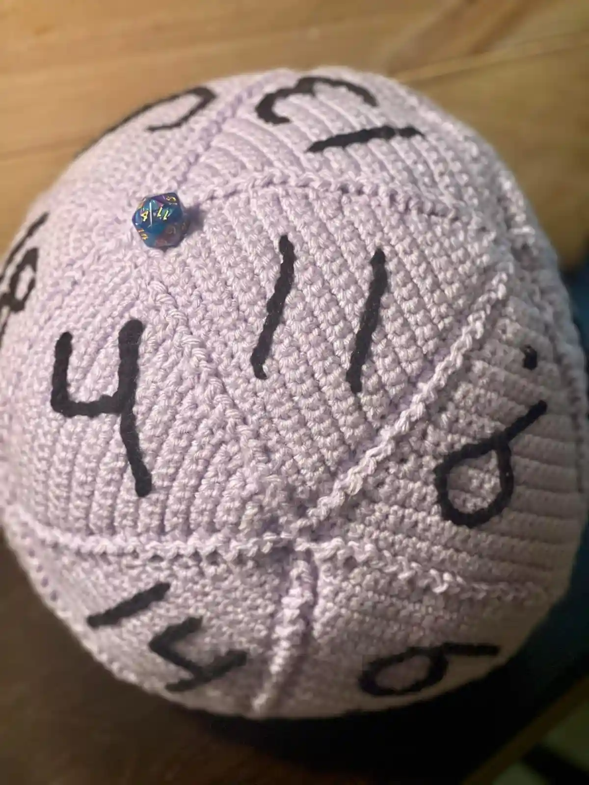A light purple crocheted d20 die cover with black numbers drawn on it, showing 4, 11, and 8. A small blue and purple…