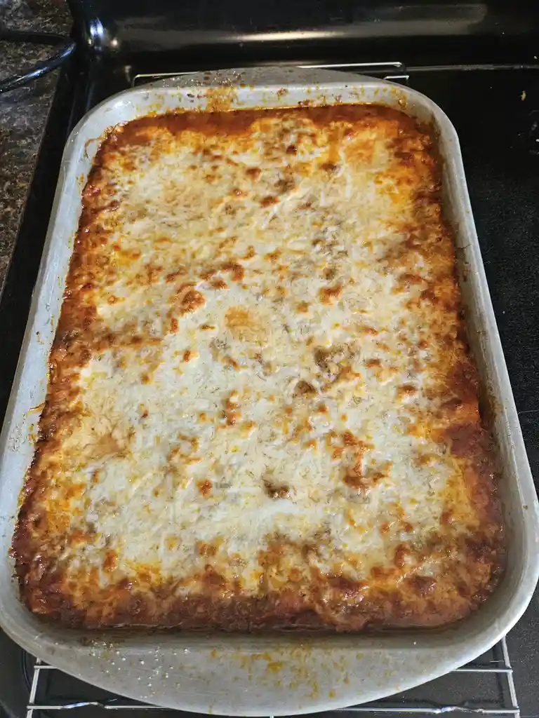 A rectangular metal baking pan sits on a cooling rack, containing a baked lasagna with a golden-brown, bubbly cheese topping…