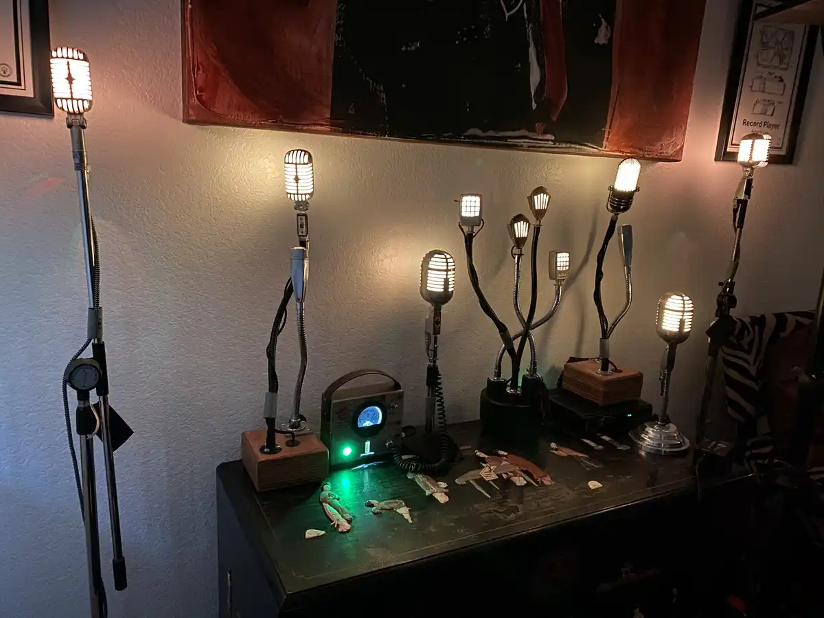 A dark room features a desk with multiple vintage-style microphones repurposed as lamps, glowing warmly. A small green-lit…