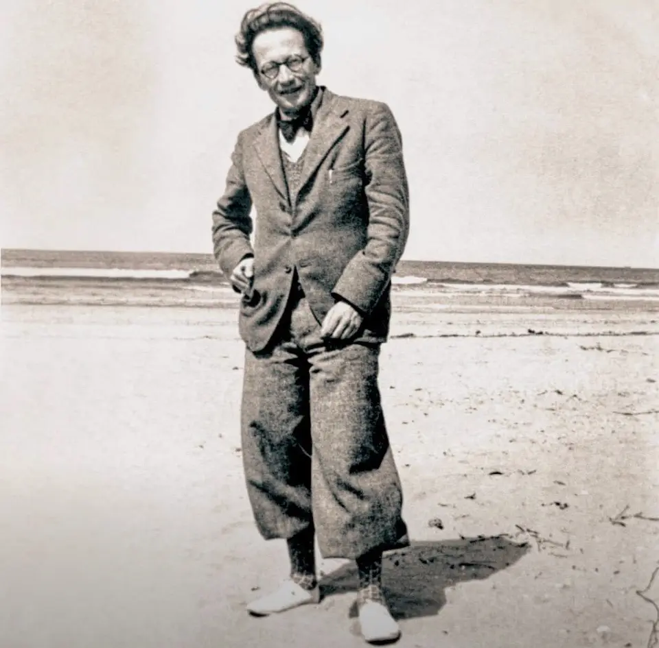 A vintage black-and-white photo shows a man with glasses and a bowtie standing on a beach, smiling while wearing loose…