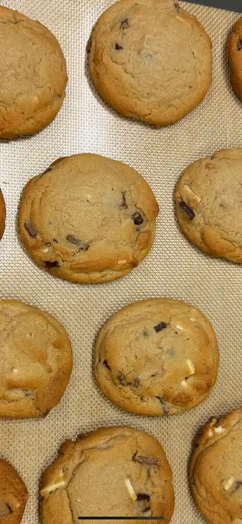 A close-up photo shows several freshly baked chocolate chip cookies arranged on a light-colored silicone baking mat. The…