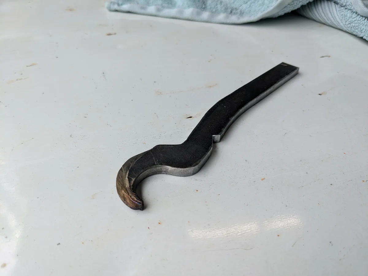 A custom-made metal wrench with a curved, mustache-like shape lies on a white work surface, with a light blue towel visible…