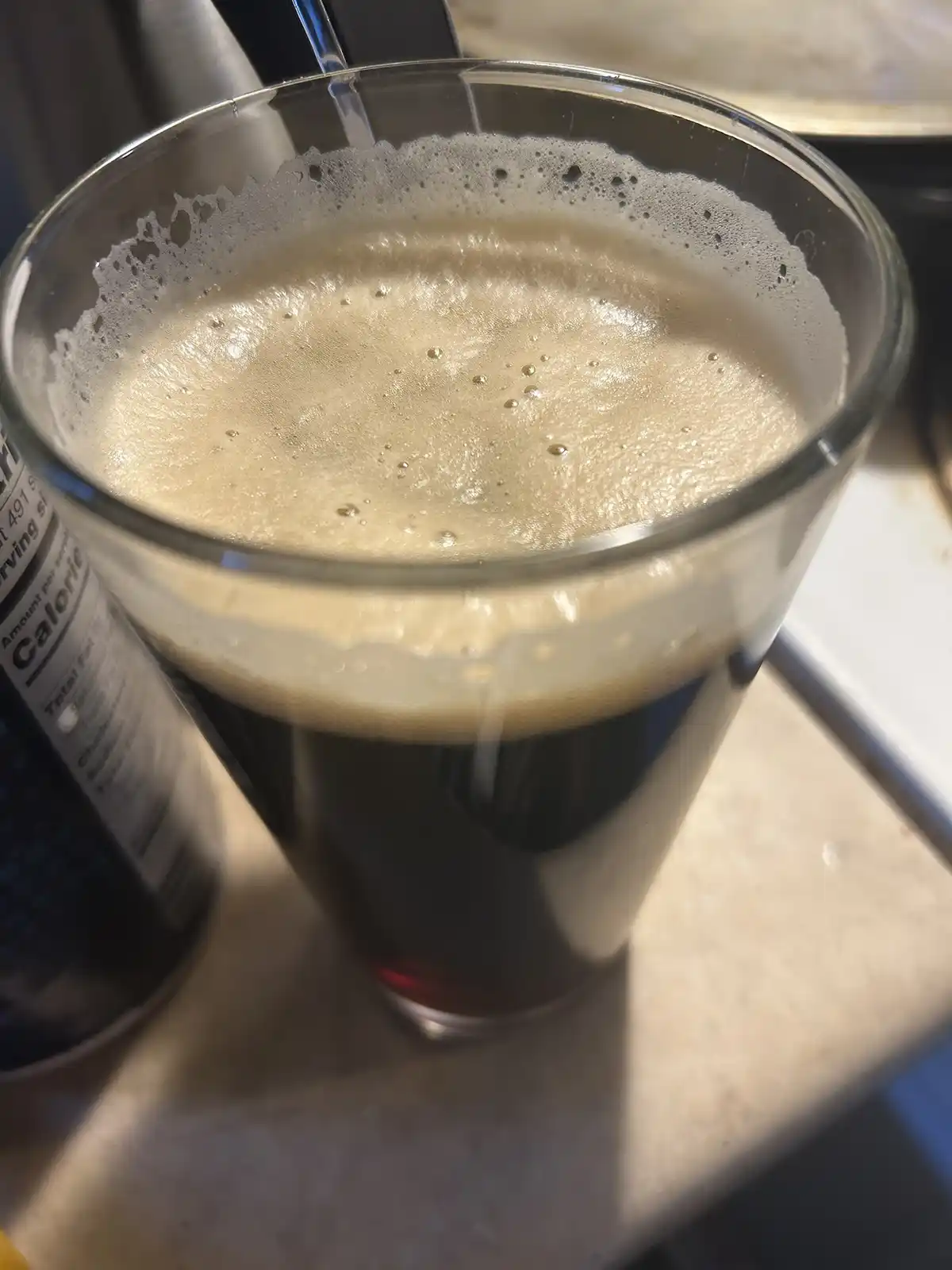 A close-up photo shows a glass mug filled with a dark beverage topped with a thick, frothy, beige head that has overflowed…