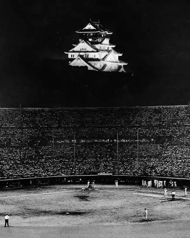 A black-and-white photo shows a baseball game at night in a large stadium filled with spectators. Dominating the dark sky…