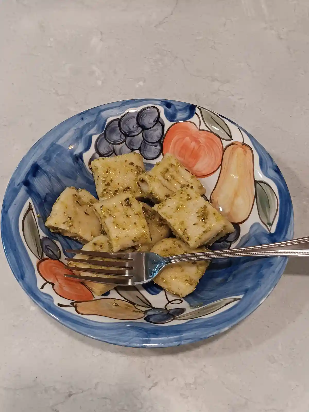 A plate with a fruit and vegetable pattern, holding cubed, pesto-coated food resembling pasta, with a fork resting on it…