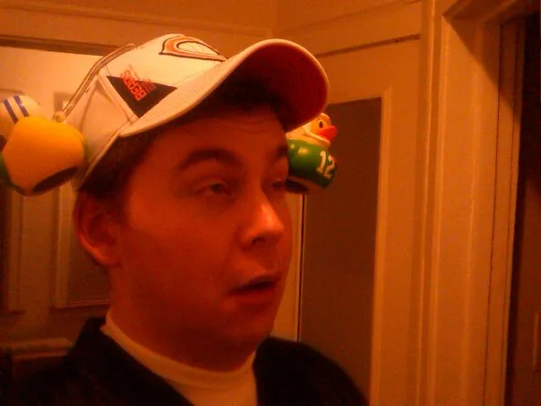 A man wearing a white baseball cap with rubber duck toys attached to the sides, one yellow and one green with the number 12…