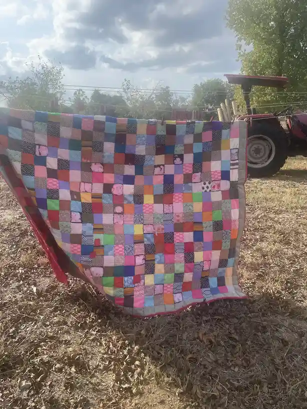 A colorful patchwork quilt hangs on a clothesline outdoors, with a red tractor visible in the background on dry grass under…