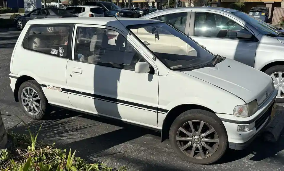 A white, slightly dirty, older model hatchback car with a black side stripe and 'pochette' written on the door, parked in a…