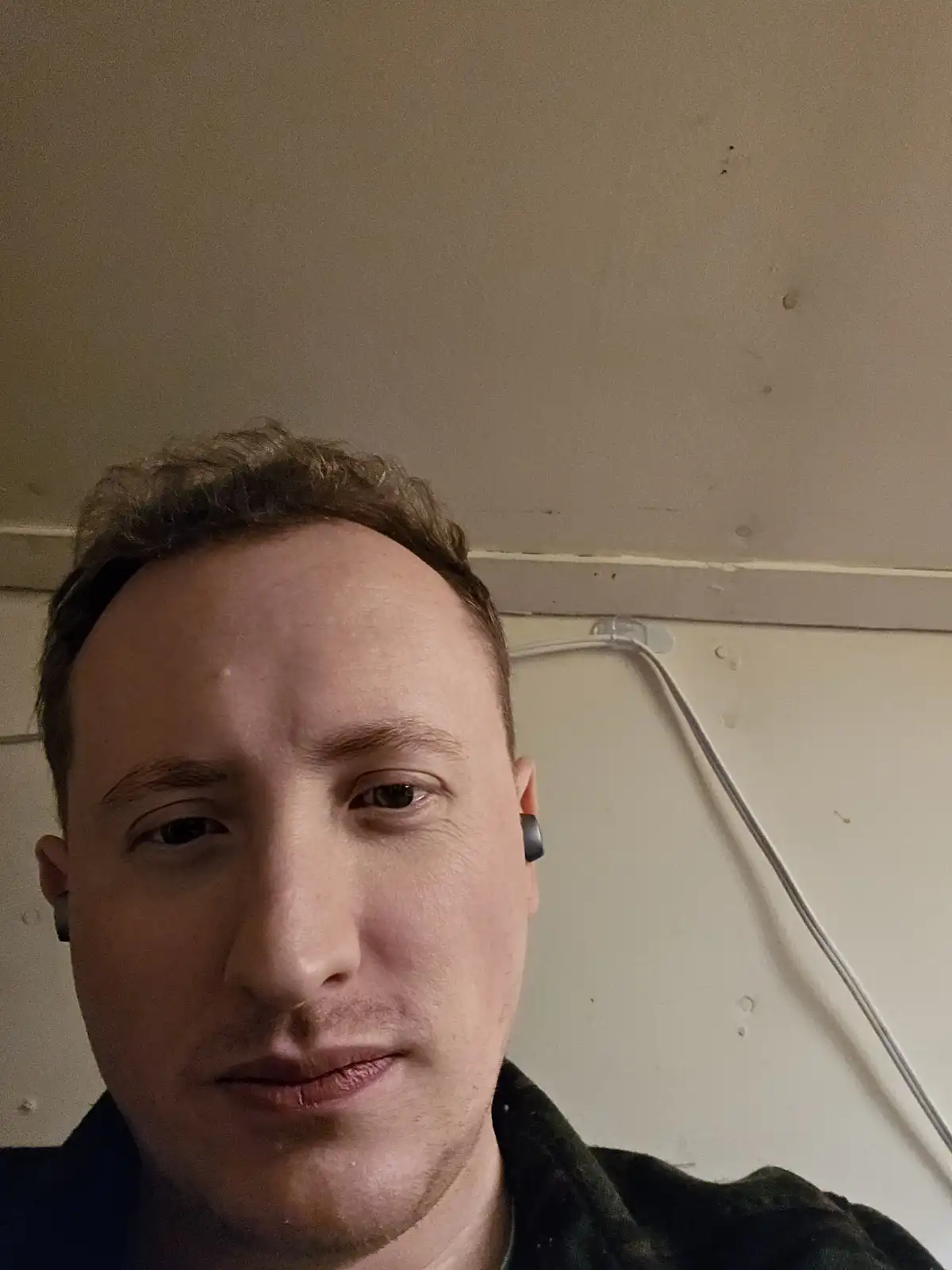 A close-up selfie of a man with short brown hair and a neutral expression, wearing black earbuds, against a plain off-white…