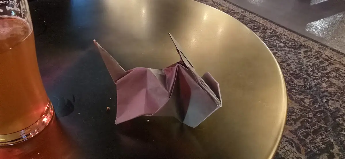 A pink origami crane sits on a gold-colored plate next to a glass of amber-colored beer on a dark table. The overlaid text…