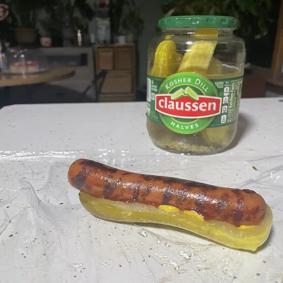 A grilled hot dog with ketchup and mustard is placed inside a dill pickle half, sitting on plastic wrap on a white…