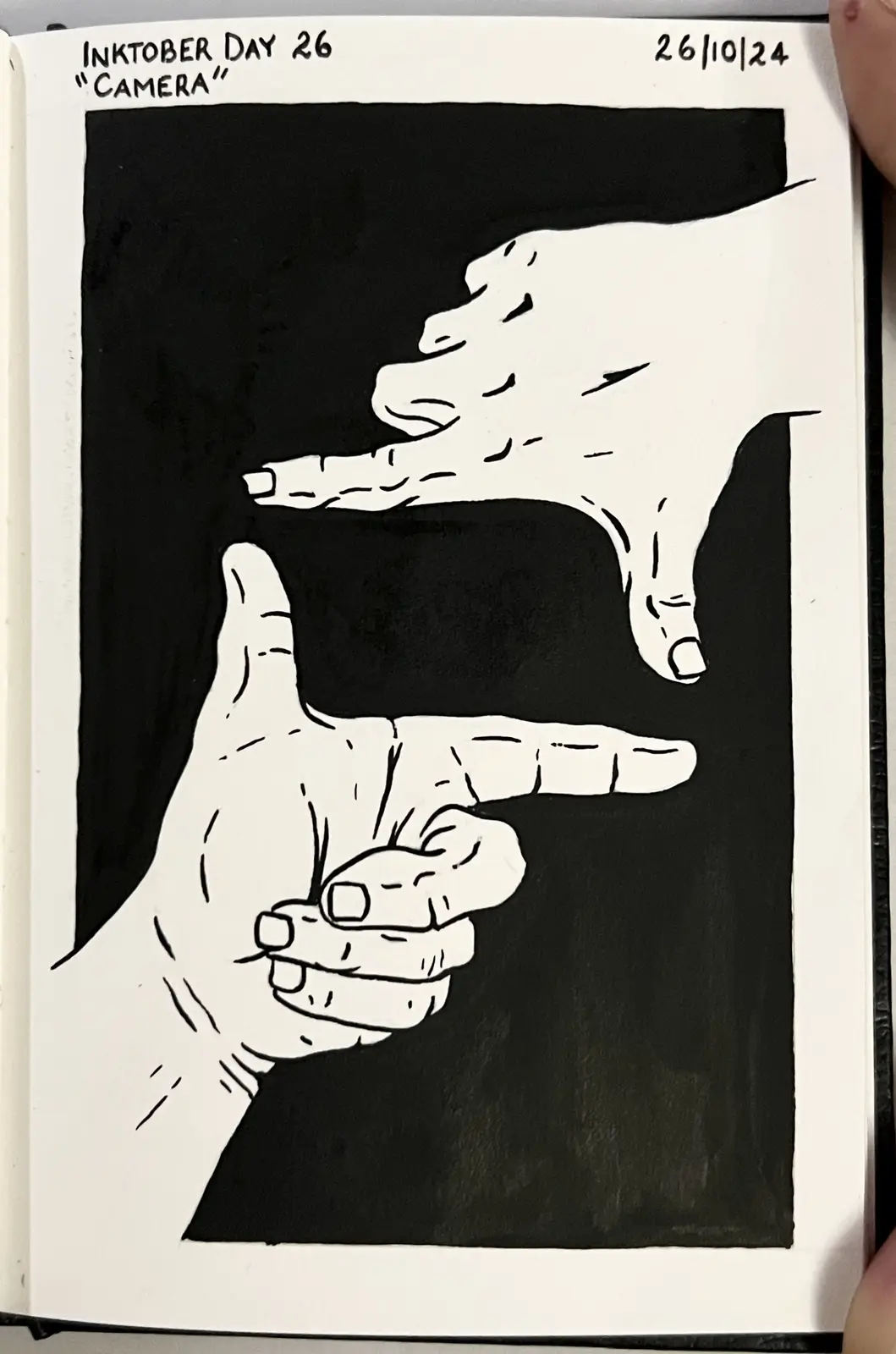 A black-and-white ink drawing in a sketchbook shows two hands forming a camera frame gesture against a black background. The…