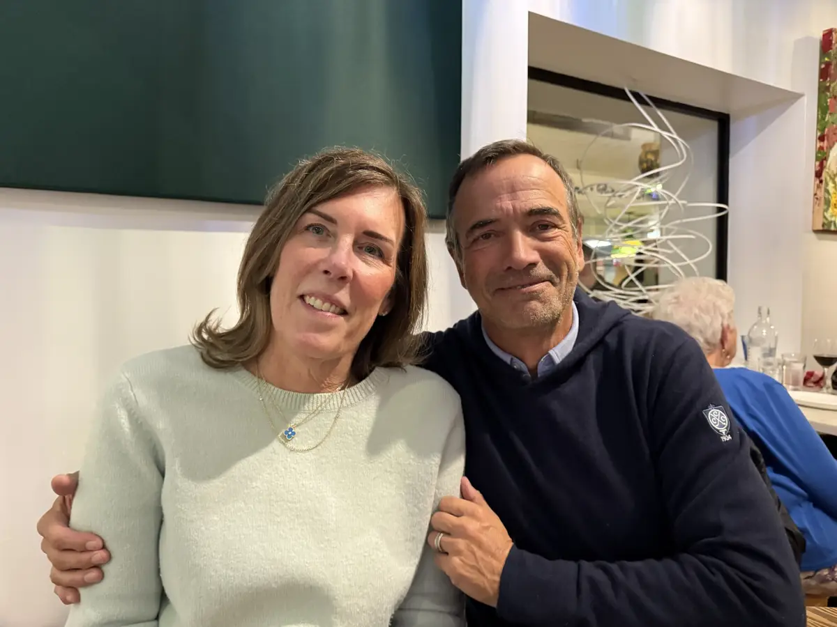 A man and woman are posing for a photo together indoors, smiling at the camera. The man has his arms around the woman's…