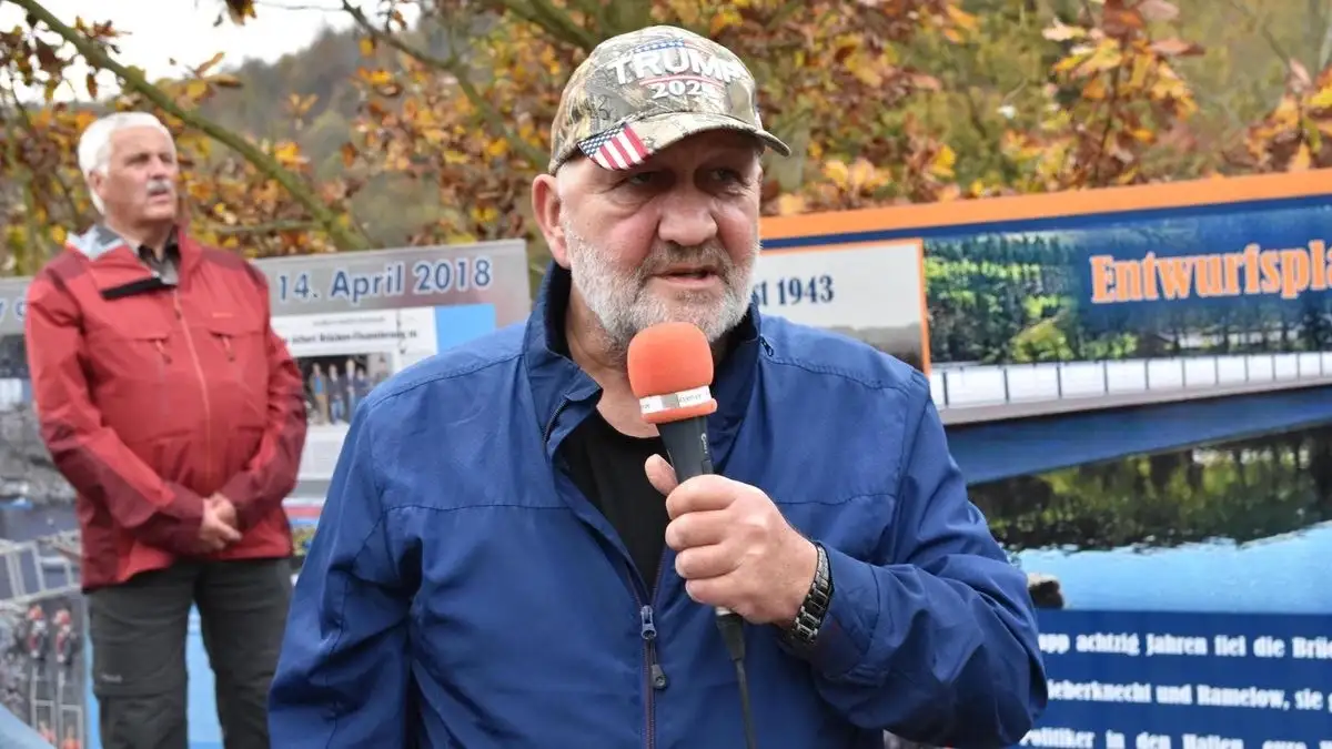 A man with a gray beard wearing a camouflage baseball cap with 'TRUMP 2025' and an American flag patch speaks into a red…