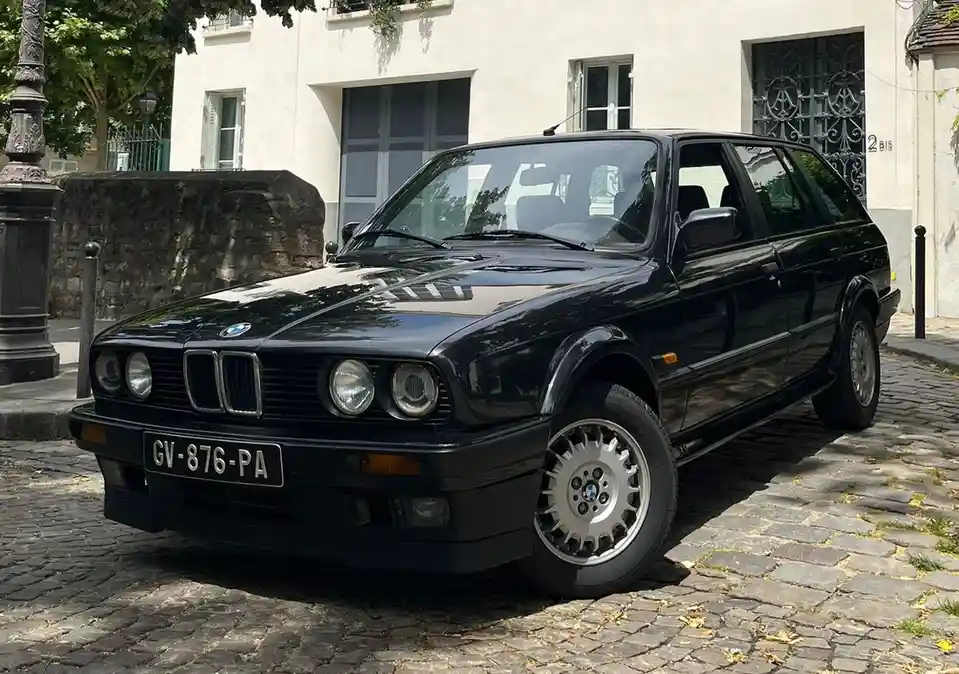 A black BMW E30 station wagon is parked on a cobblestone street in front of a white building. The car has a glossy finish…