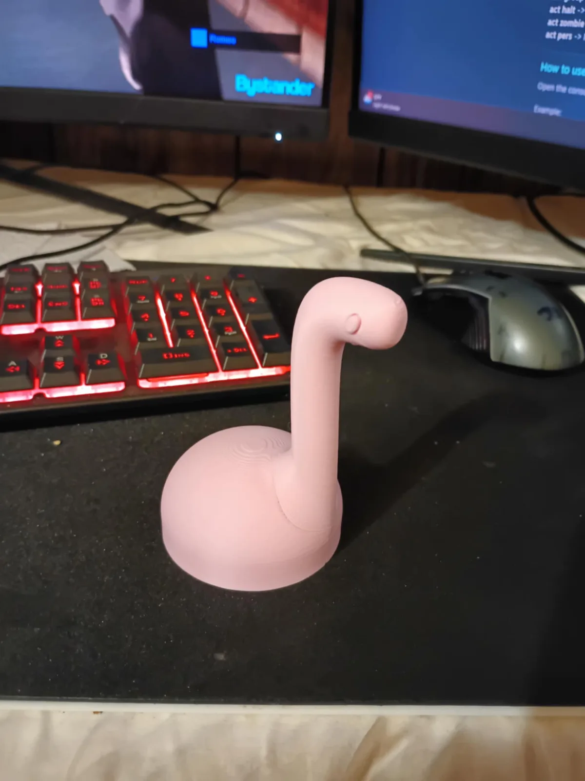 A pink, 3D-printed dinosaur-like figurine with a long neck sits on a black desk in front of a red-backlit keyboard and two…