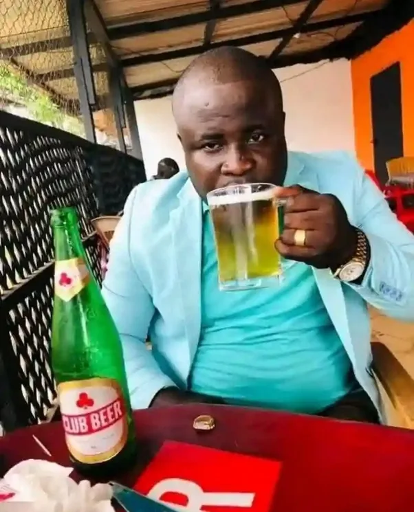 a man in a light blue suit is drinking from a glass mug while sitting at a red table with a green bottle of Club Beer beside…