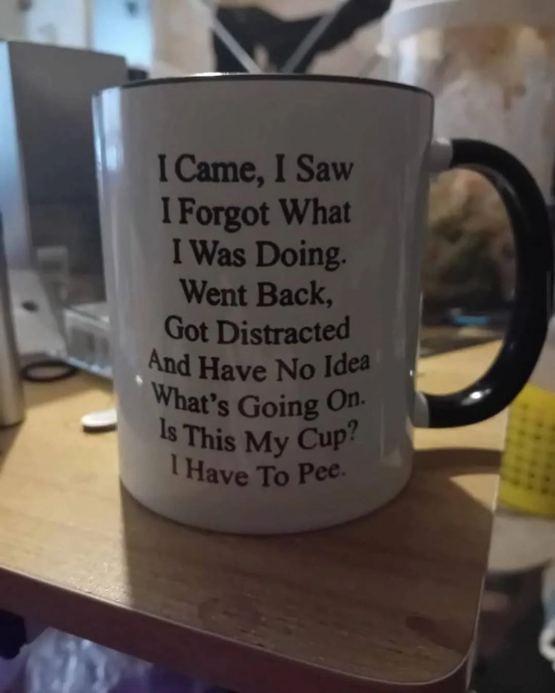 A white ceramic mug with a black handle sits on a wooden desk, featuring a humorous text that reads: 'I Came, I Saw I Forgot…