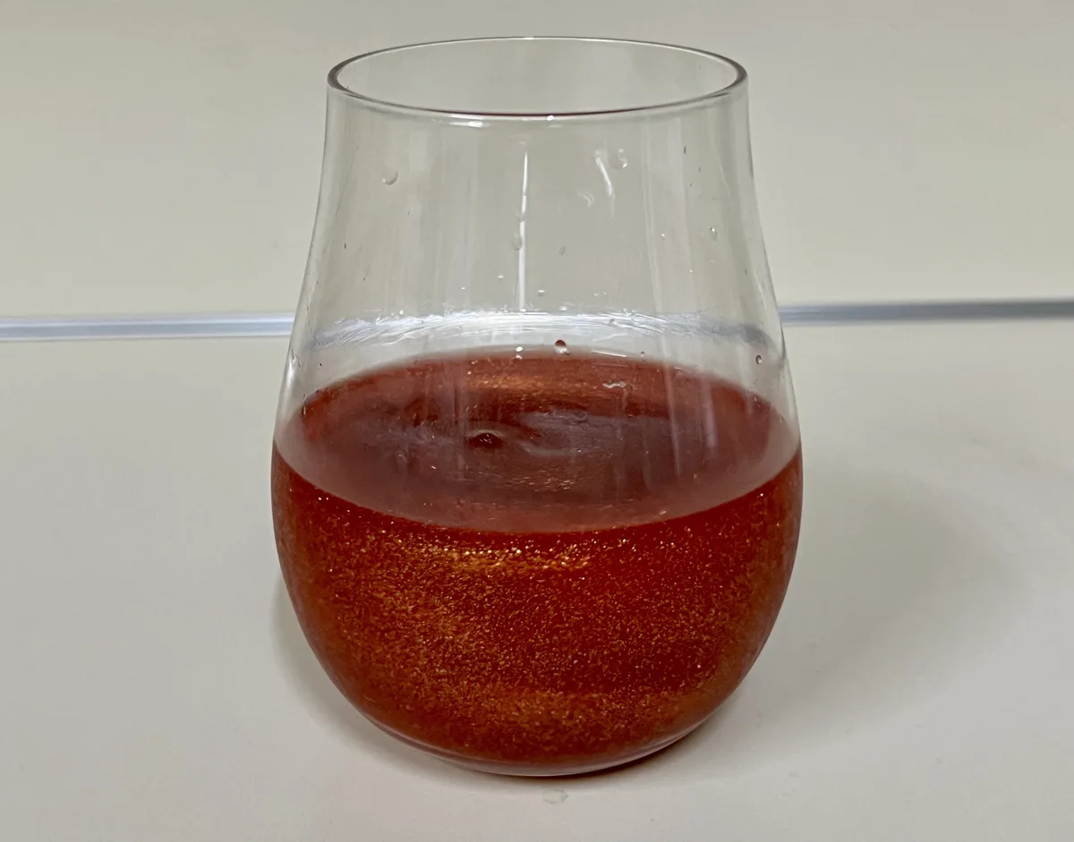 A clear glass containing a reddish-orange liquid with visible glittery particles sits on a white surface. The overlaid text…