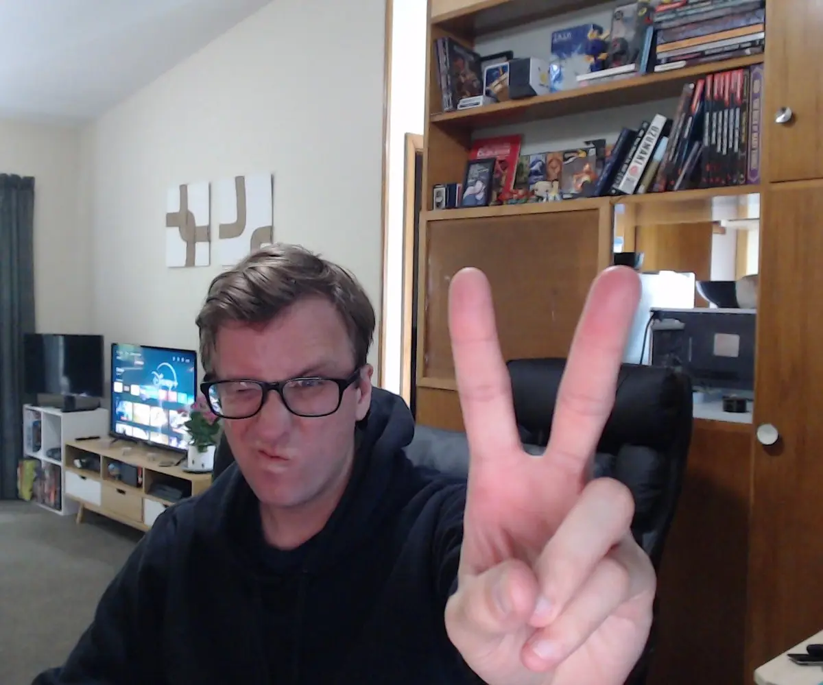 A man wearing glasses and a black hoodie sits in a chair in a living room, making a peace sign with his hand while making a…