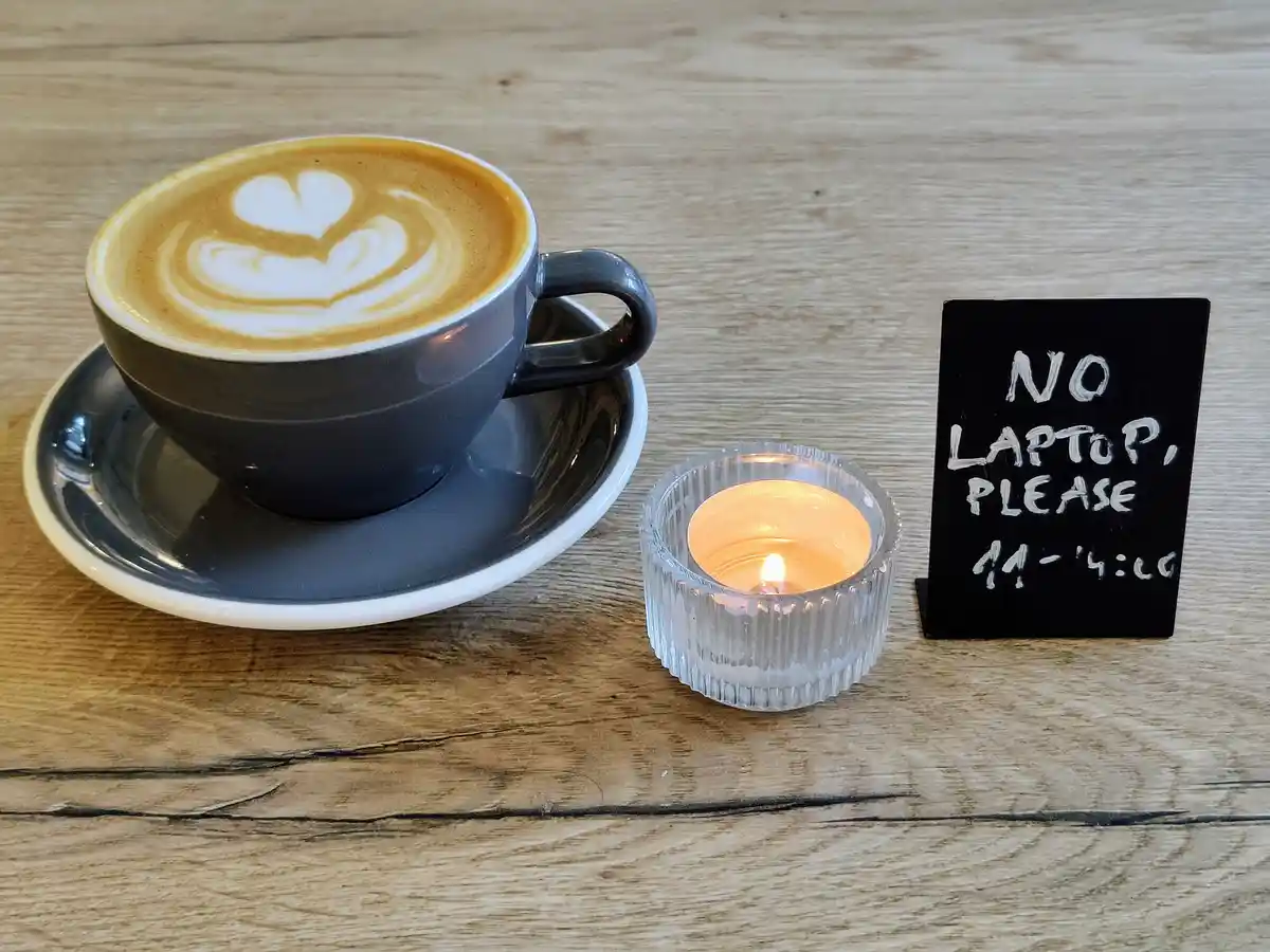 A dark grey coffee cup with heart-shaped latte art sits on a saucer next to a lit candle in a glass holder and a small black…