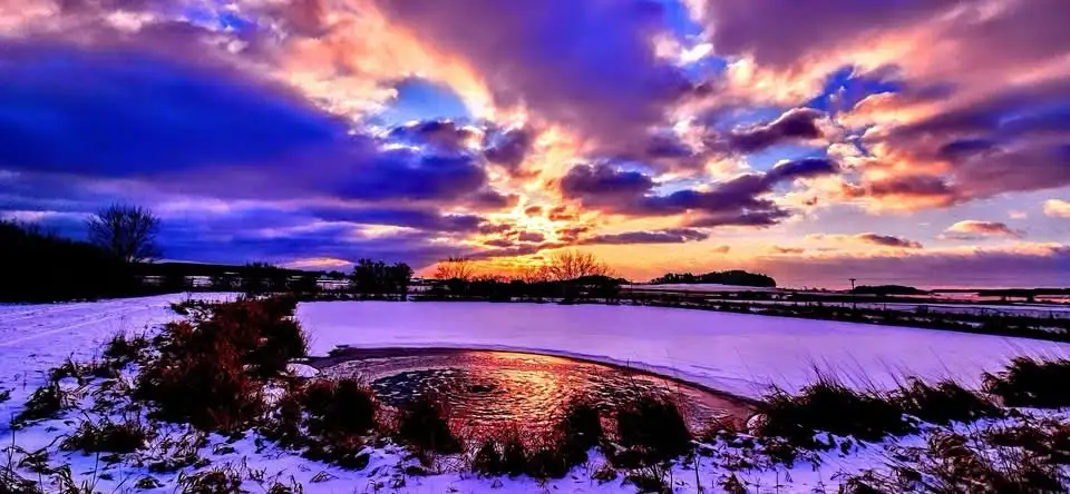 A snowy landscape at sunset with vibrant purple and orange clouds reflecting in a partially frozen pond. The overlaid text…