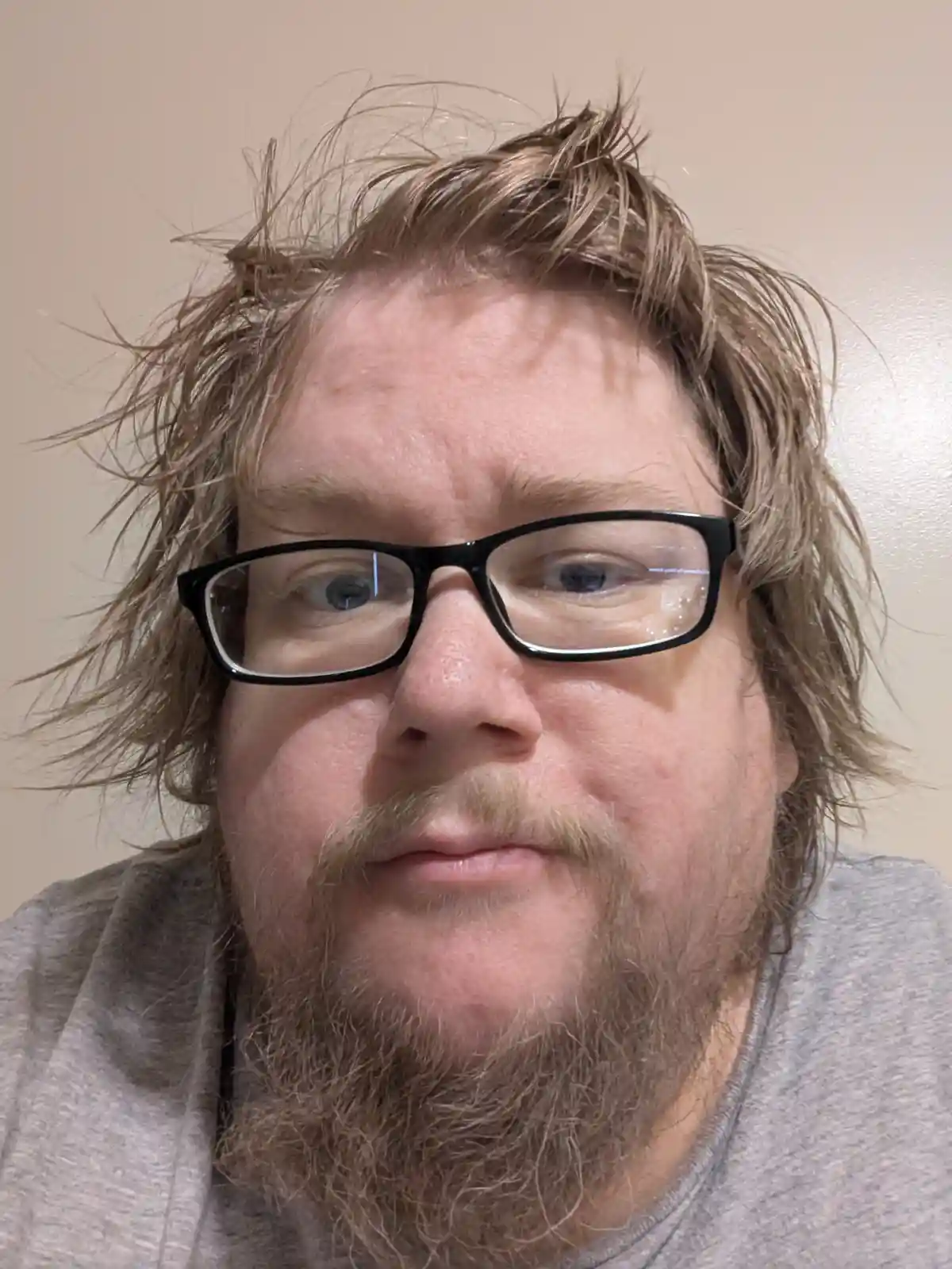 A close-up photo of a man with messy, light brown hair, wearing black-rimmed glasses and a full beard, looking directly at…