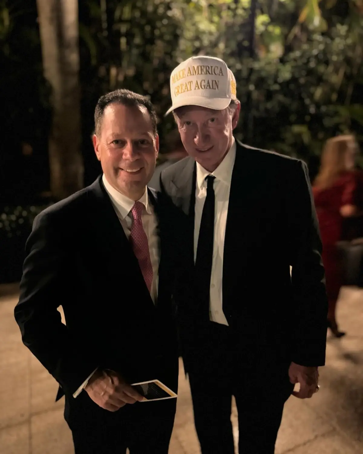 Two men in suits pose for a photo at night outdoors with trees in the background. The man on the right wears a white…