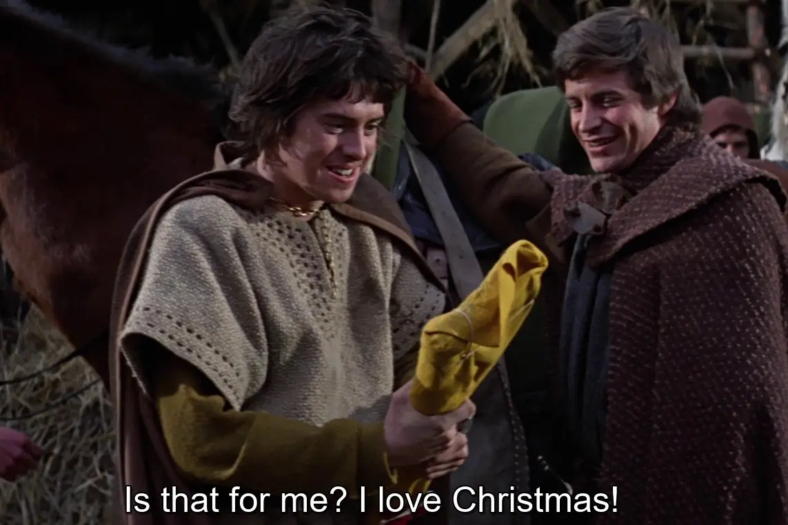 Two men in medieval-style clothing stand together; the man on the left, holding a yellow cloth bundle, smiles broadly while…