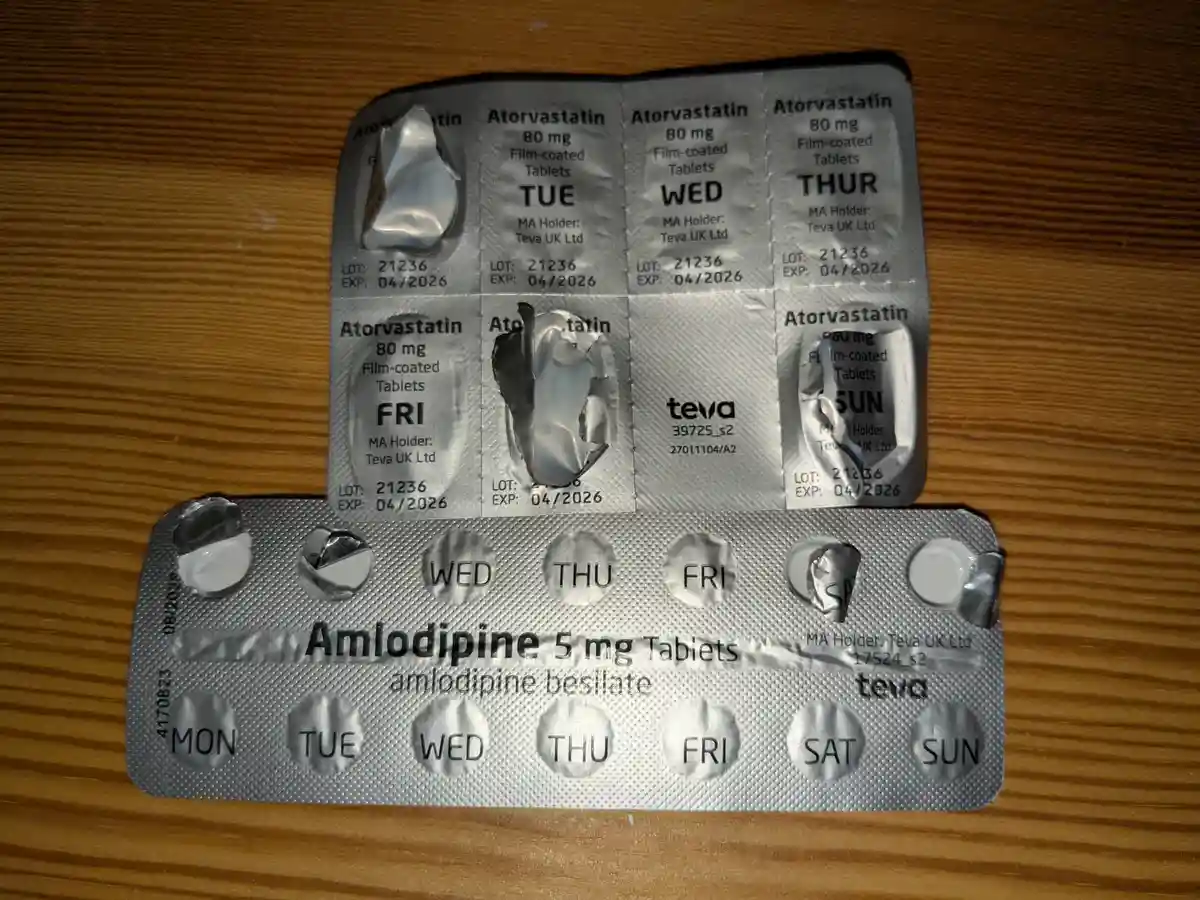 Two blister packs of prescription medication lie on a wooden surface. The top pack contains Atorvastatin 80mg tablets…