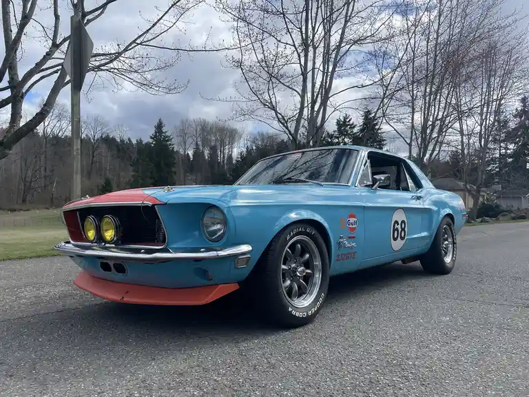 A light blue 1968 Ford Mustang with red accents and racing decals, including a Gulf Oil logo and the number 68 on its side…