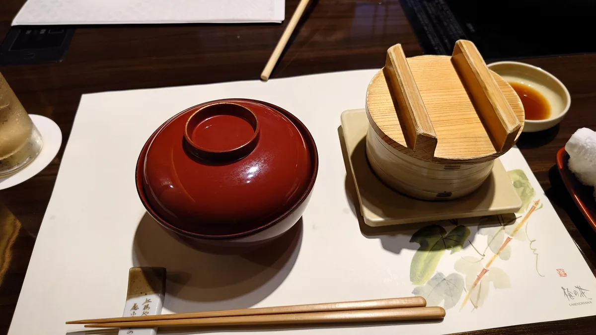 a close-up photo of a Japanese restaurant table setting featuring a glossy red lacquer lidded bowl, a bamboo steamer with a…