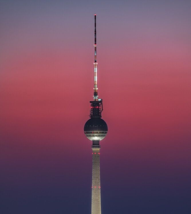 A tall, slender tower with a spherical observation deck and antenna array stands against a gradient sky transitioning from…