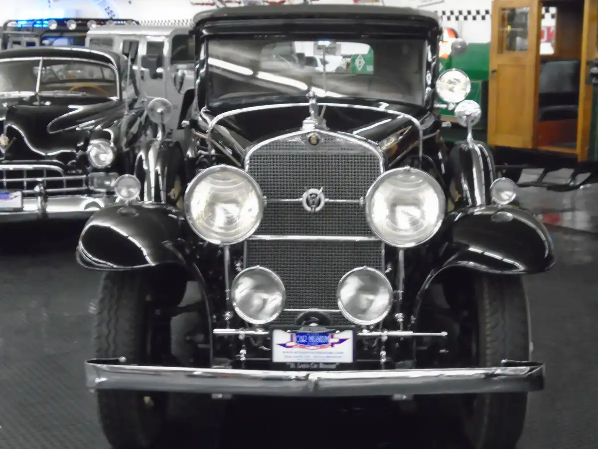 A close-up frontal view of a shiny black vintage car with large round headlights and chrome accents, displayed indoors among…
