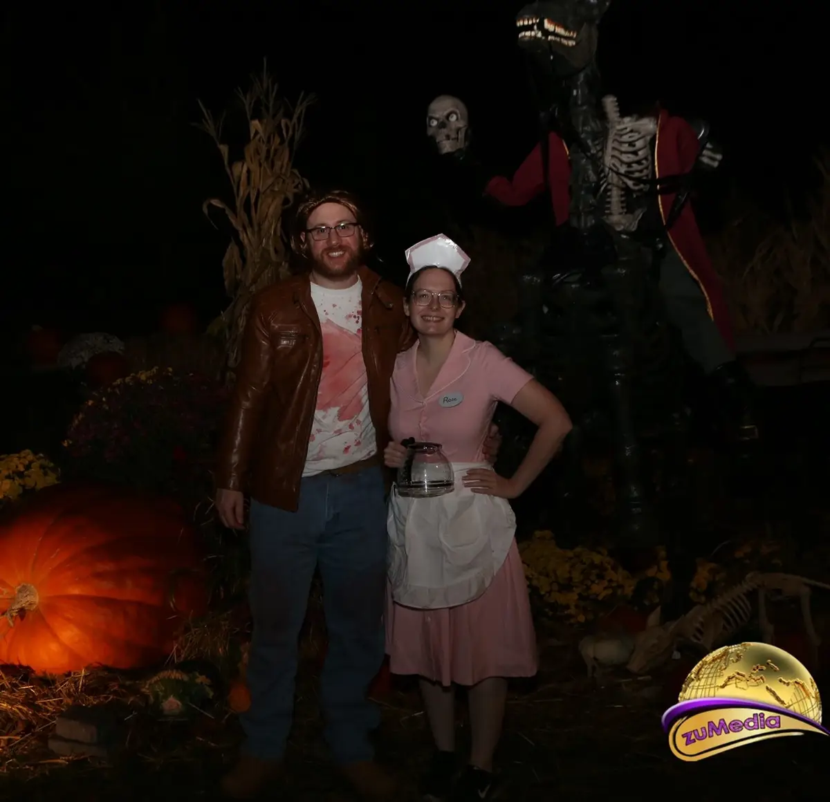 Two people pose for a photo at a Halloween-themed outdoor setting with pumpkins, cornstalks, and spooky props like a…